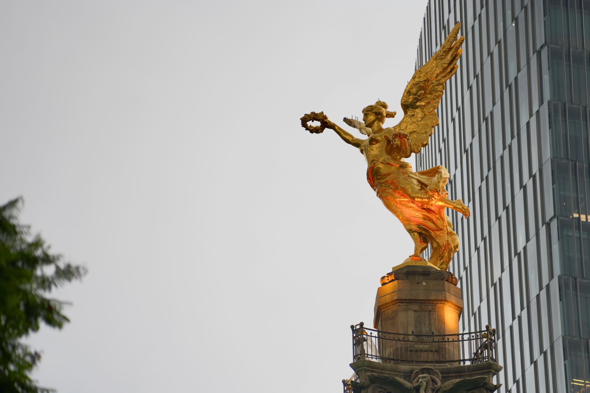 El Ángel de la Independencia de día, dorado contra un cielo gris y torres de cristal, la corona en alto.