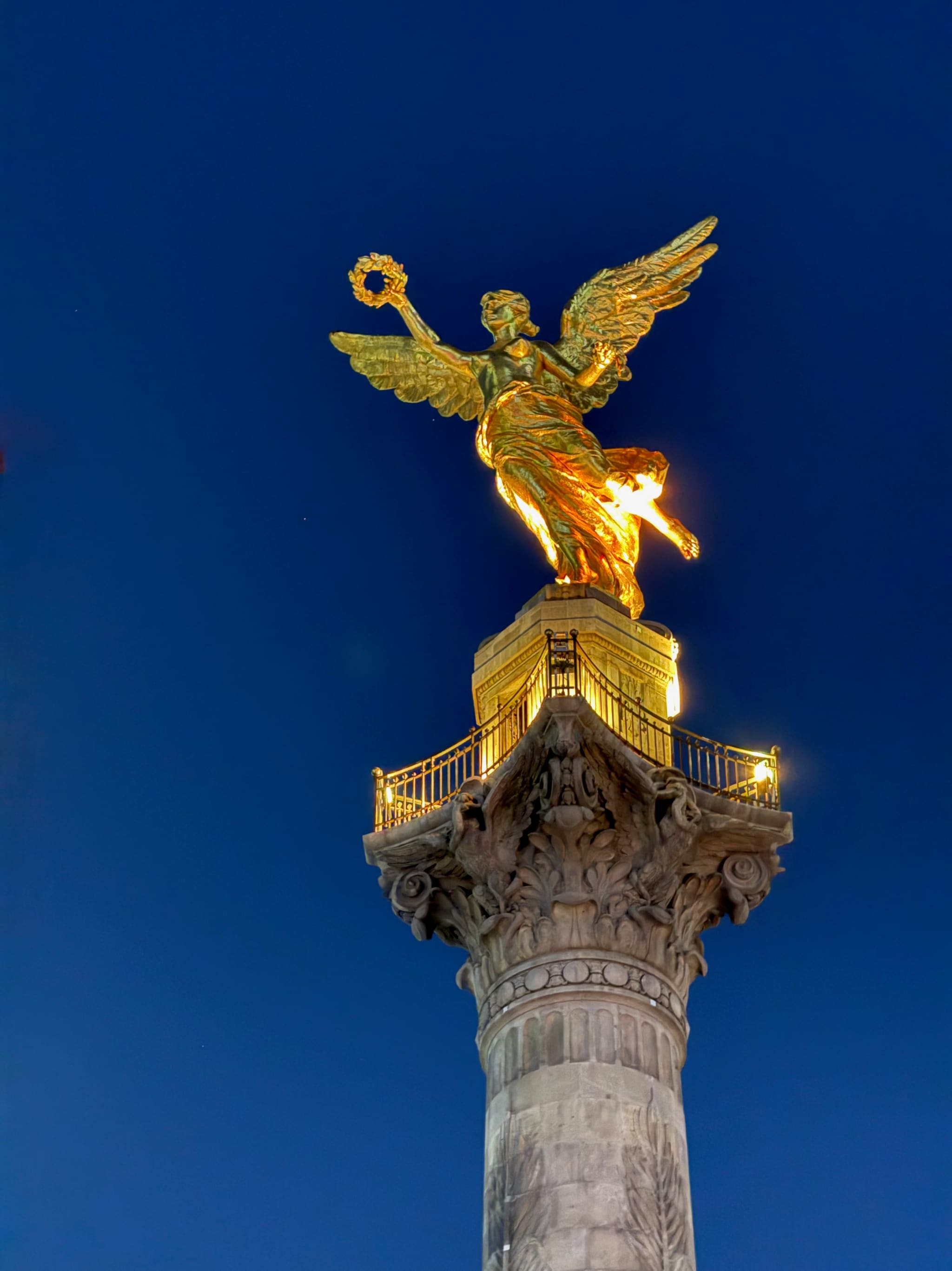 El Ángel de la Independencia de noche, bronce dorado iluminado contra un cielo azul profundo, la corona en alto.