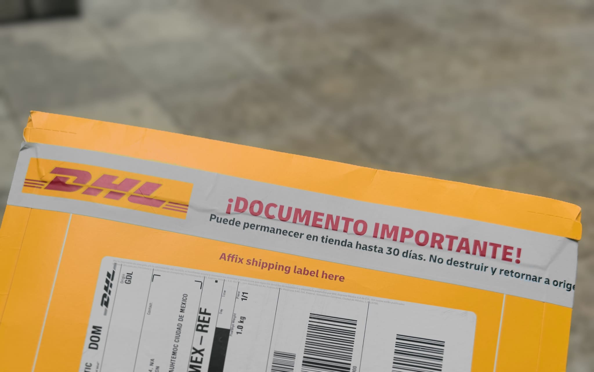 The top of a yellow DHL courier envelope, its cardboard pull-tab still sealed, photographed over blurred stone. A grey band carries red print: '¡DOCUMENTO IMPORTANTE!' — and beneath it, 'Puede permanecer en tienda hasta 30 días. No destruir y retornar a origen': it may remain in the store up to thirty days; do not destroy, return to origin.