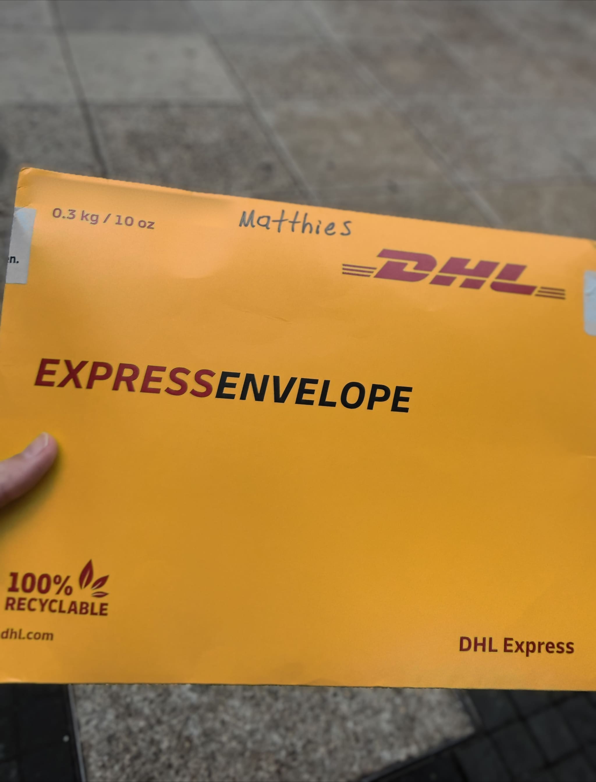 A hand holds a yellow DHL Express envelope over wet pavement. 'Matthies' is handwritten at the top beside the printed weight — 0.3 kg / 10 oz — and the words EXPRESS ENVELOPE run across the middle.