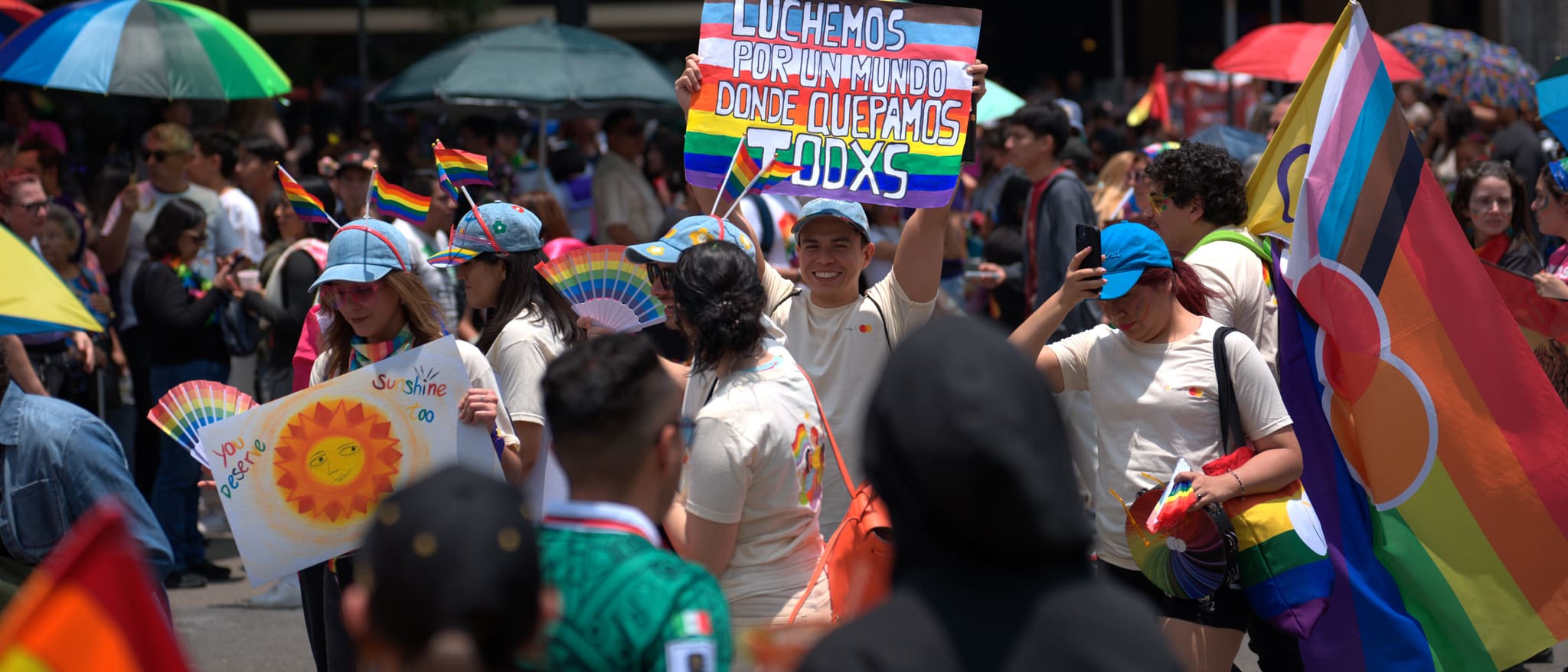 Una multitud densa del Orgullo bajo sombrillas arcoíris en una avenida de la Ciudad de México; al centro, un letrero pintado a mano con las franjas de la bandera del progreso: «Luchemos por un mundo donde quepamos todxs».