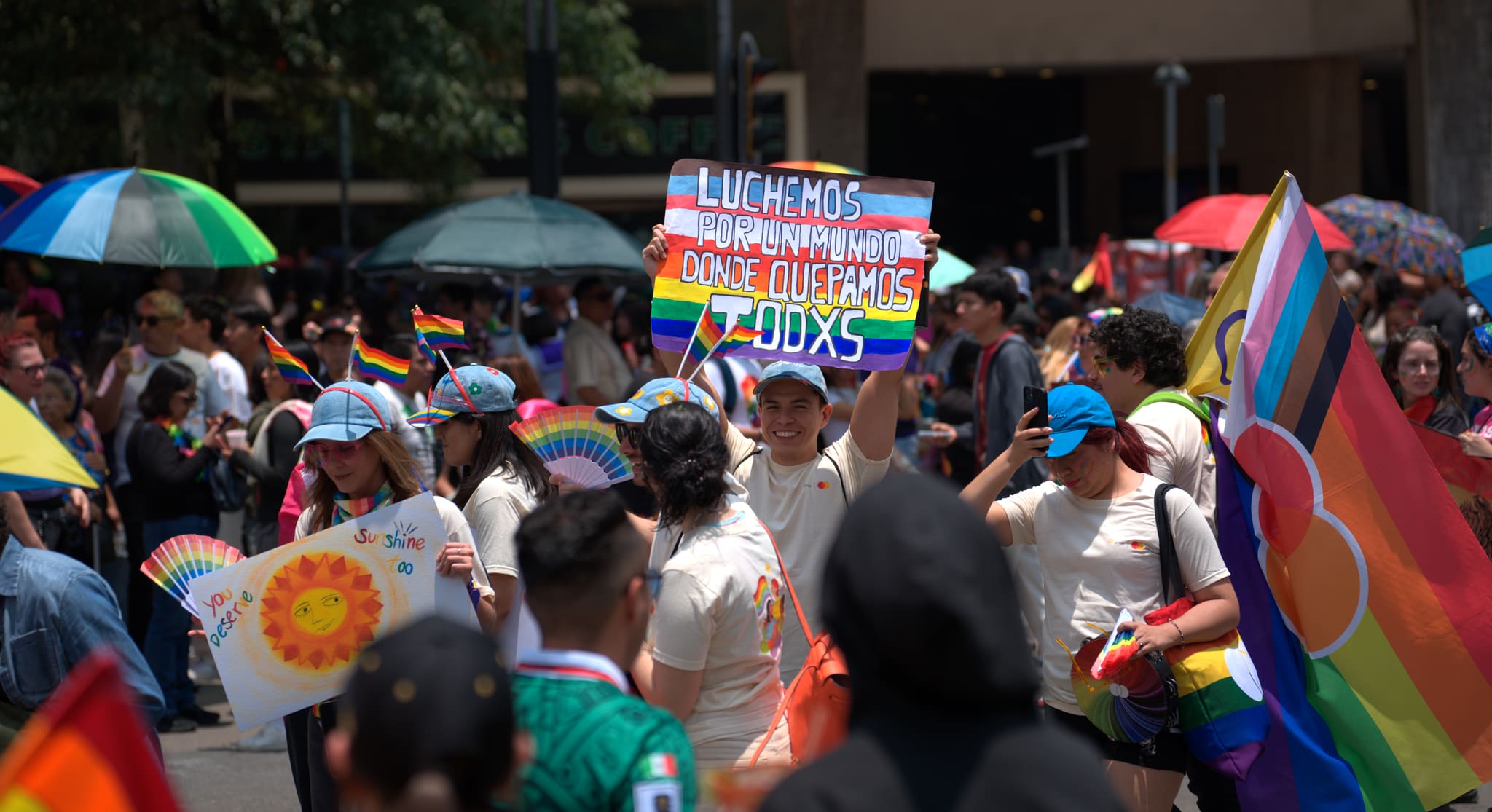 Una multitud densa del Orgullo bajo sombrillas arcoíris; al centro, un letrero pintado a mano con las franjas de la bandera del progreso: «Luchemos por un mundo donde quepamos todxs».