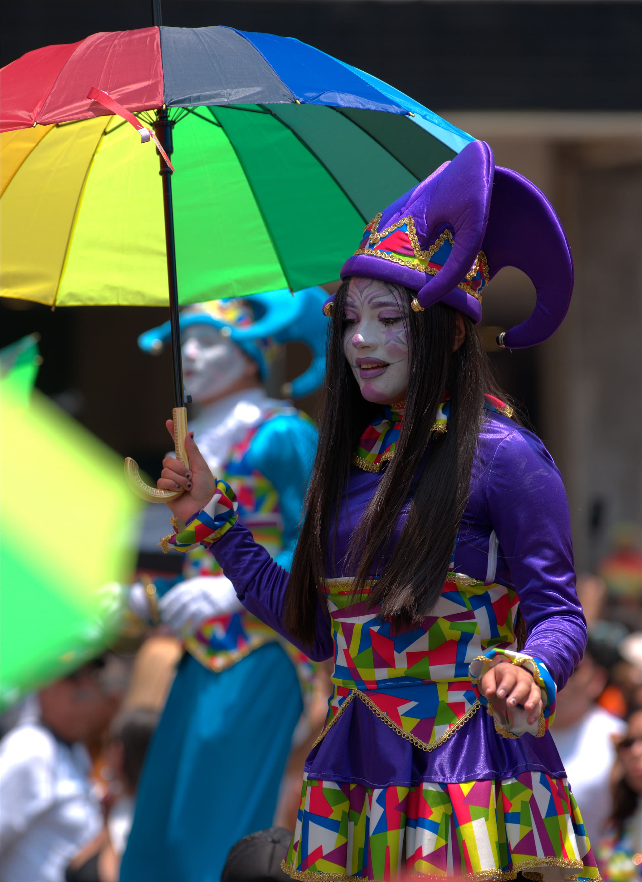 Una artista con traje de bufón morado y la cara pintada de blanco bajo una sombrilla arcoíris, los ojos bajos, a medio paso.