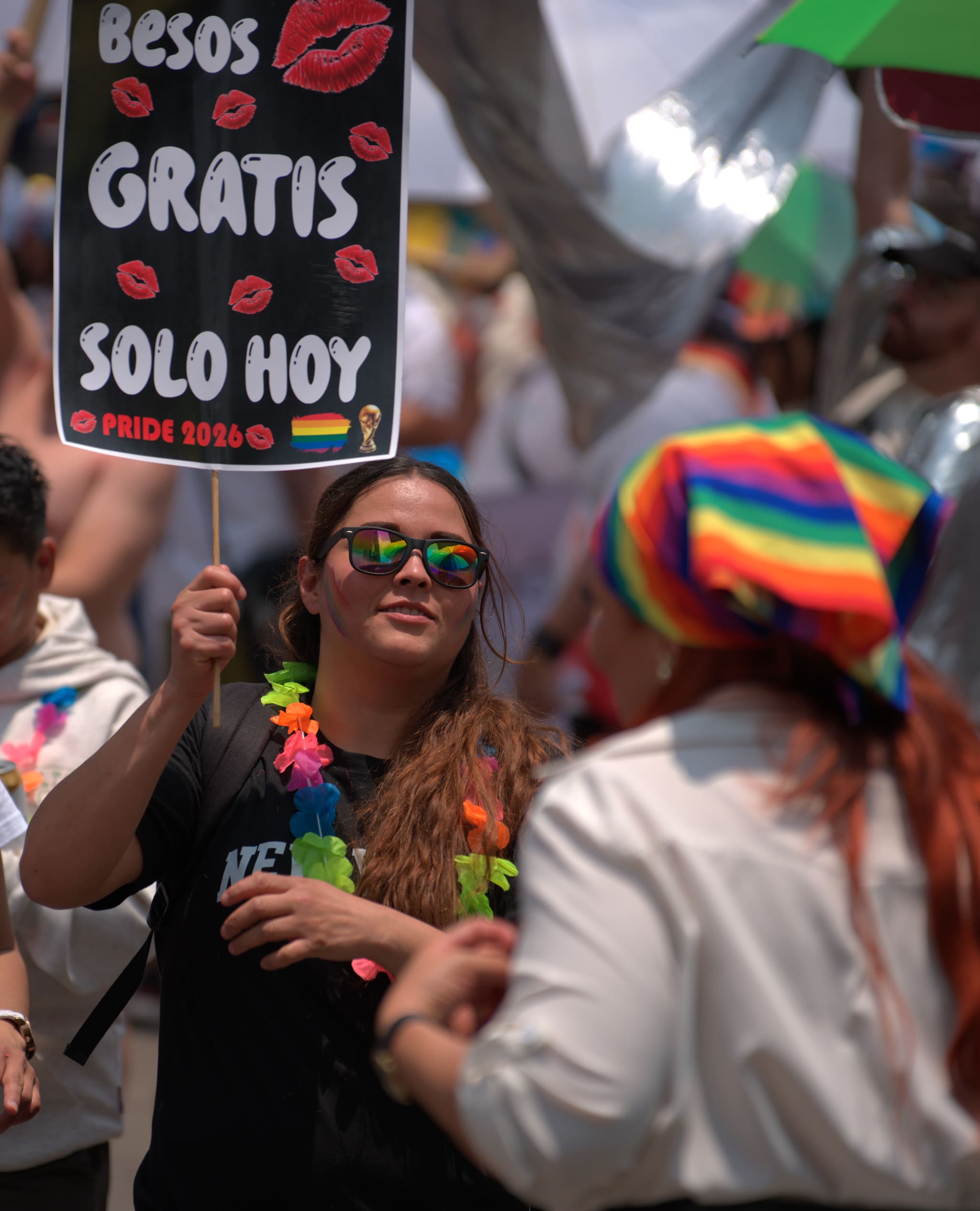 Una mujer sonriente con lentes de espejo arcoíris sostiene una pancarta negra que dice «Besos gratis, solo hoy — Pride 2026», con una bandera arcoíris y la copa del Mundial impresas en la esquina.