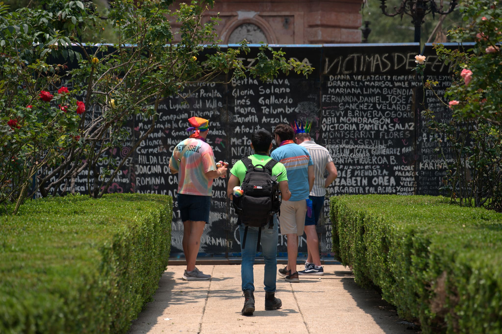 Cuatro personas con colores del Orgullo leen, de pie, un largo muro negro cubierto de nombres escritos a mano con gis — víctimas de feminicidio — en un rosedal.