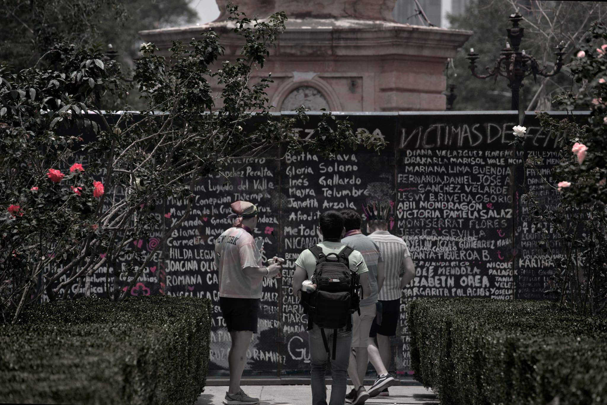 Four men stand reading a long, hand-chalked memorial wall at the back of a rose garden, beneath a header that begins 'VÍCTIMAS DE FEM—'. Dozens of women's names fill the panels — Mariana Lima Buendía plainly legible — with hearts drawn beside some. The photograph is drained nearly to monochrome: only the roses, the pink chalk marks, and a faint blush on the monument behind keep their color.