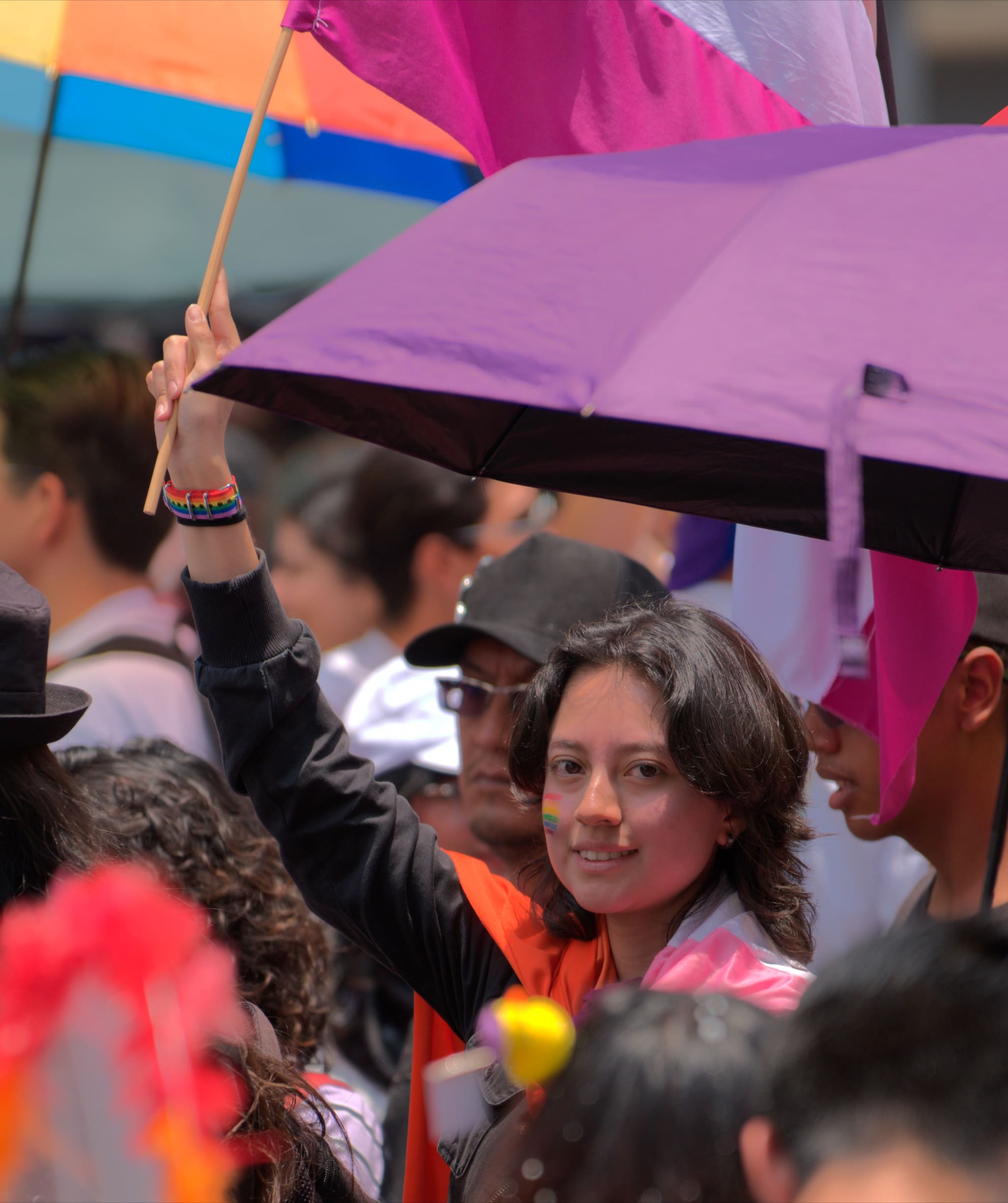 Una persona bajo una sombrilla morada en una multitud del Orgullo, con un pequeño arcoíris en la mejilla, mirando hacia la cámara.